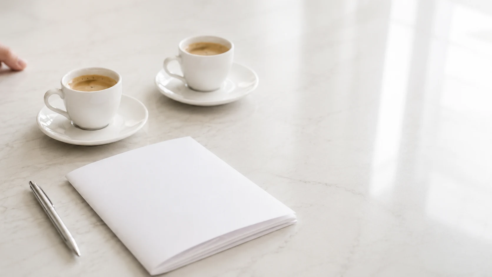 Two espresso cups beside a folded blank document and a pen on a marble counter